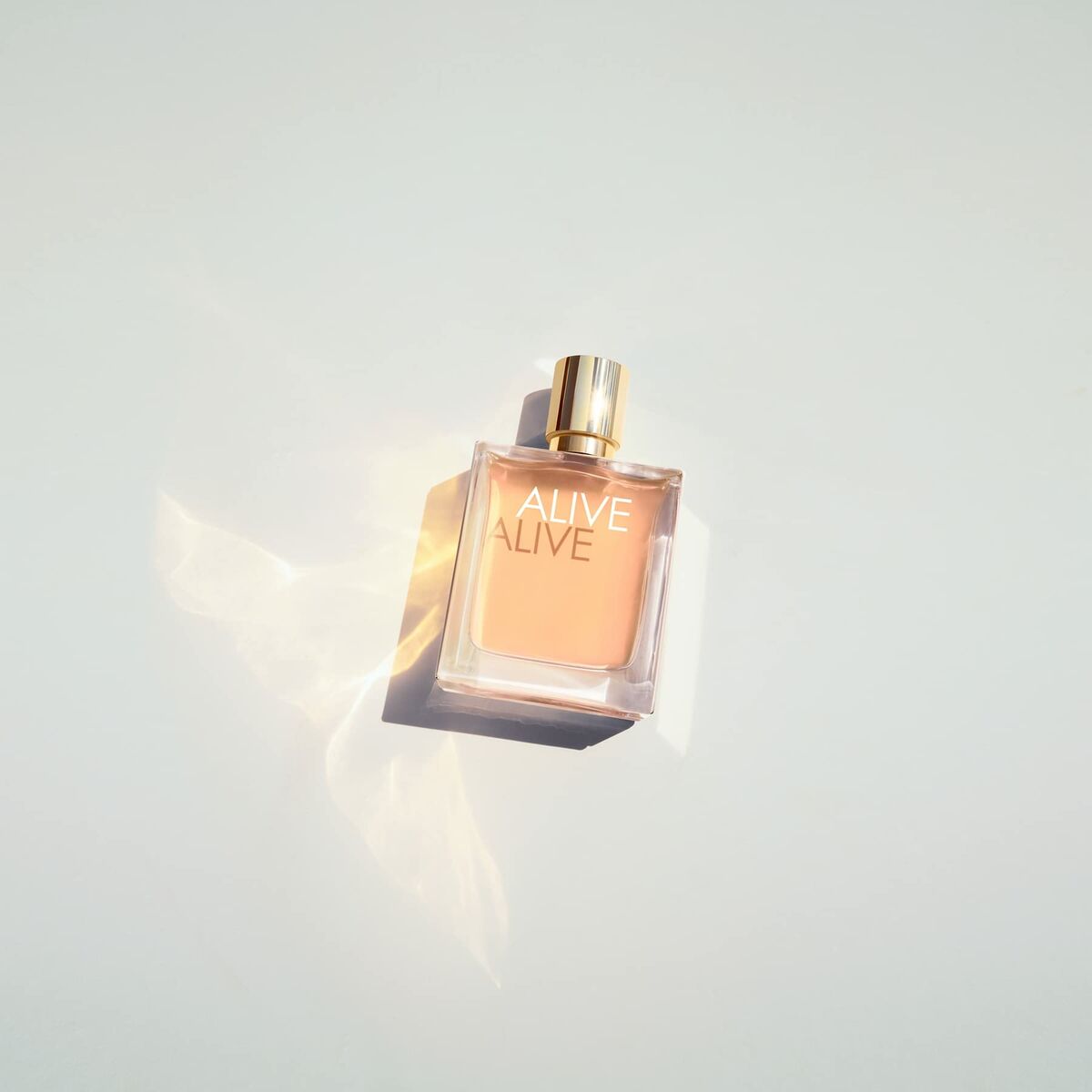 Flacon de parfum 'ALIVE' avec étiquette dorée, posé sur une surface blanche brillante, rayons de lumière créant un effet d'ombre.