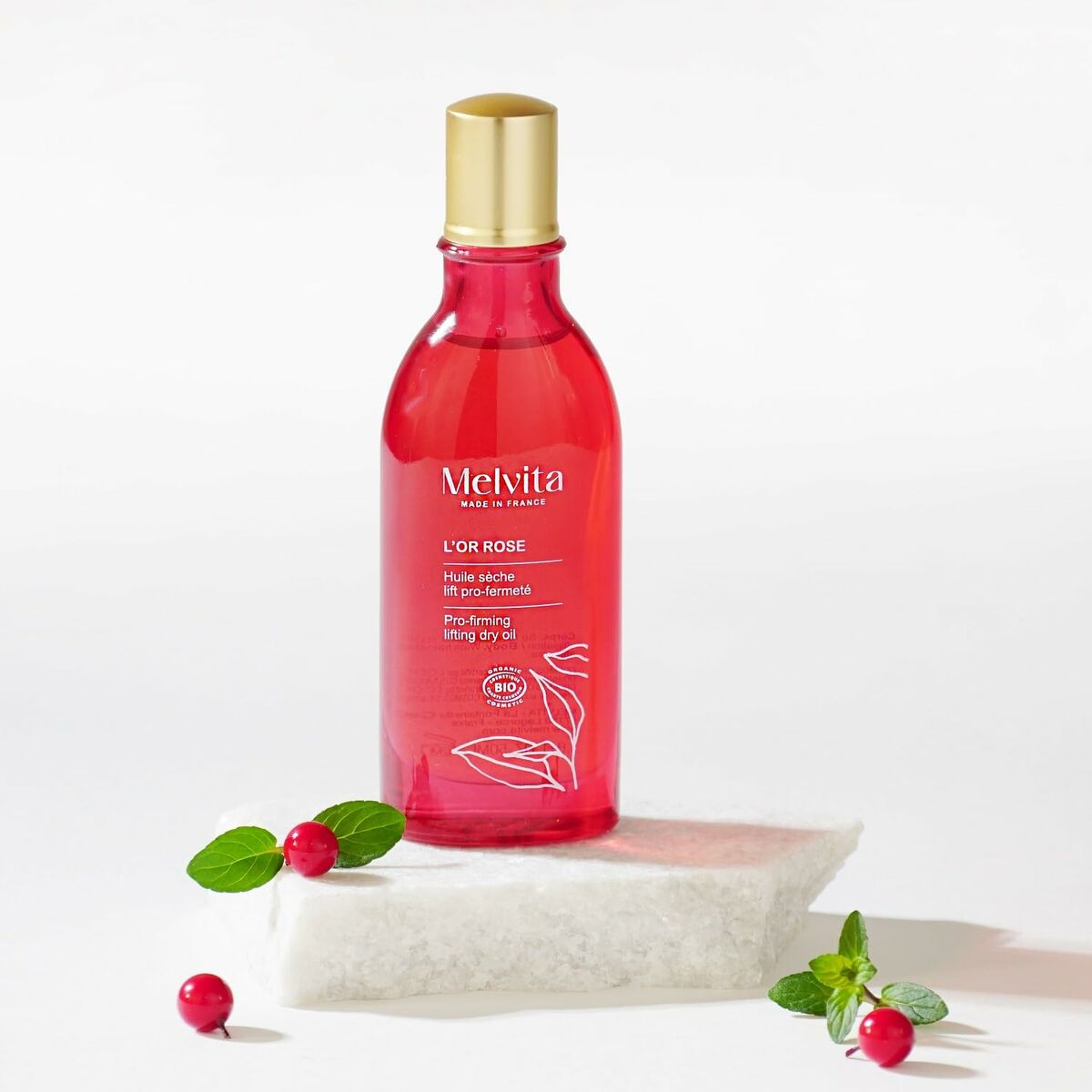 Lotion corporelle Melvita L'OR ROSE, flacon rouge avec bouchon doré, posé sur un support blanc avec des baies et feuilles.