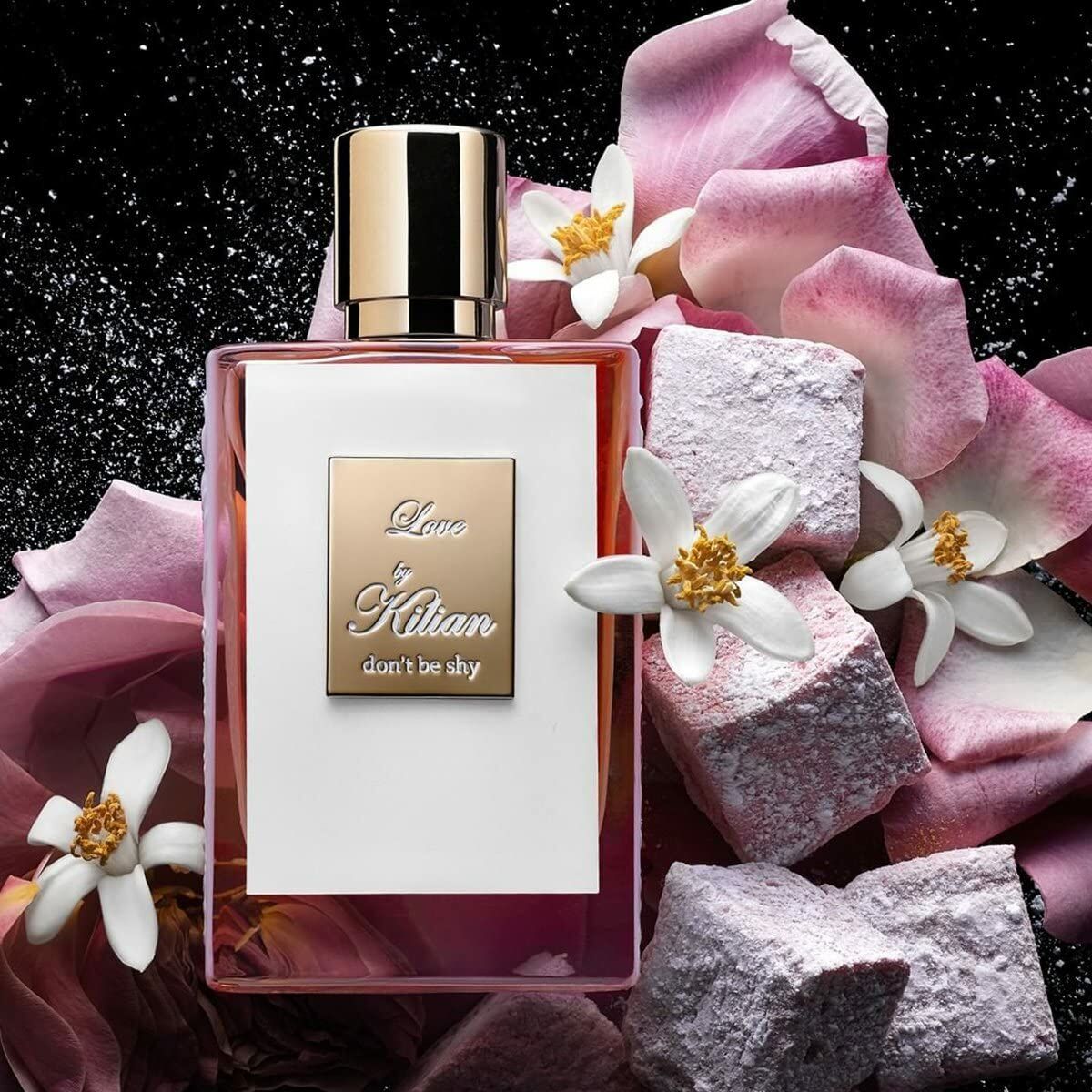 Kilian Love Don't Be Shy EDP 50 ml bottle surrounded by pink flowers and marshmallows on a dark background.