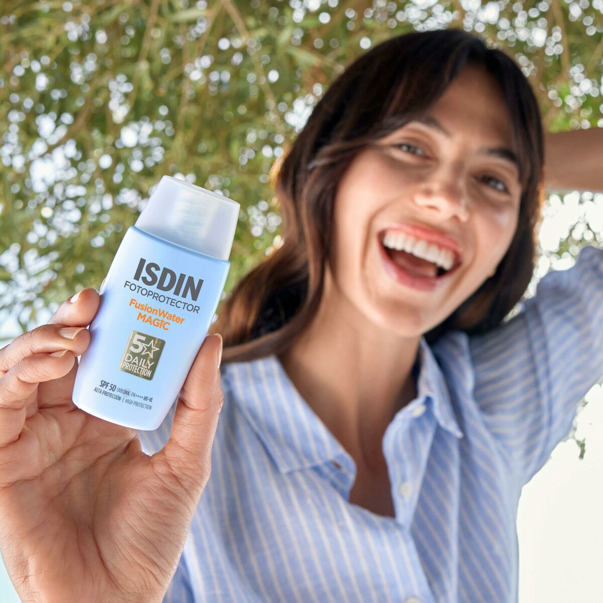 Femme souriante montrant le Protecteur Solaire Isdin FOTOPROTECTOR sous un arbre.